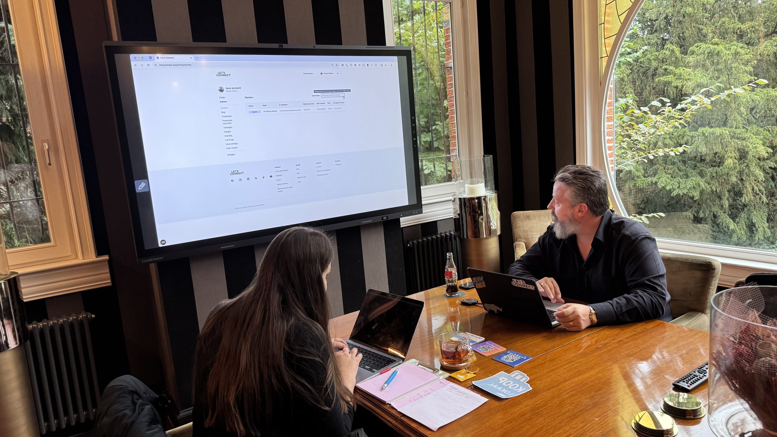 Twee mensen werken aan laptops tijdens een vergadering met schermpresentatie in een modern kantoor met grote ramen.